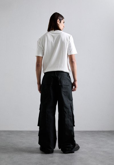 Person with long dark hair wearing a white t-shirt and black wide-leg cargo pants standing on a gray floor against a plain white wall.