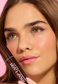 Brauner Mascara-Stift mit strukturiertem Spiraldesign, neben einem Gesicht mit glatter Haut, vollen Lippen und definierten Wimpern vor einem pinken Hintergrund gehalten.