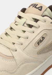 Kremfargede sneakers med overdel i semsket skinn, snøring, Fila-logo, og lysegrønn detalj. Har teksturert materiale og en brun såle.