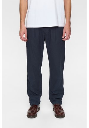 Uomo che indossa pantaloni blu navy, camicia bianca e scarpe in pelle marrone, in piedi davanti a uno sfondo bianco uniforme.