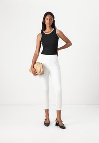 Top sin mangas negro con ribetes blancos, pantalones ajustados blancos y zapatos de tacón negros. Sosteniendo un bolso beige, con texturas suaves en toda su superficie.