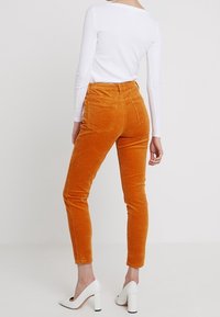 Pomarańczowe spodnie kordowe, dopasowane z tylnymi kieszeniami, w połączeniu z białą bluzką z długim rękawem i białymi szpilkami.
