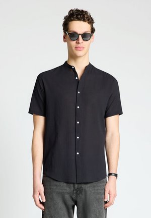 Giovane uomo che indossa una camicia nera a maniche corte con bottoni, occhiali da sole neri, jeans grigi e un orologio nero al polso, in piedi davanti a uno sfondo chiaro uniforme.