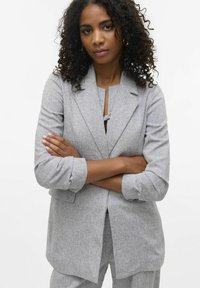 Blazer gris texturizado con cuello de solapa y mangas recogidas. El modelo tiene los brazos cruzados, vistiendo una blusa gris a juego por debajo.