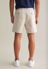 Pantalones cortos de color beige claro hechos de algodón con un acabado texturizado, que cuentan con dos bolsillos traseros y detalles de botones marrones. Combinados con zapatillas blancas.