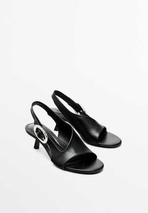 Sandales noires en cuir avec un bout ouvert, un petit talon et un accent en métal argenté en forme de cercle sur le côté. Texture lisse et design simple.