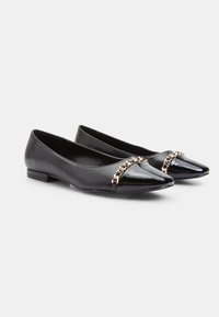 Black flat shoes made of patent leather with a pointed toe, featuring a gold chain accent across the front and a low block heel.