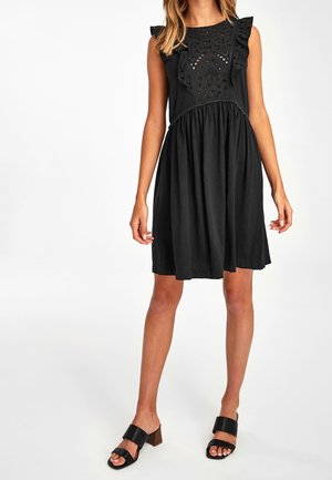 Woman wearing sleeveless black dress with eyelet details and ruffles on chest, paired with black open-toe heeled sandals.