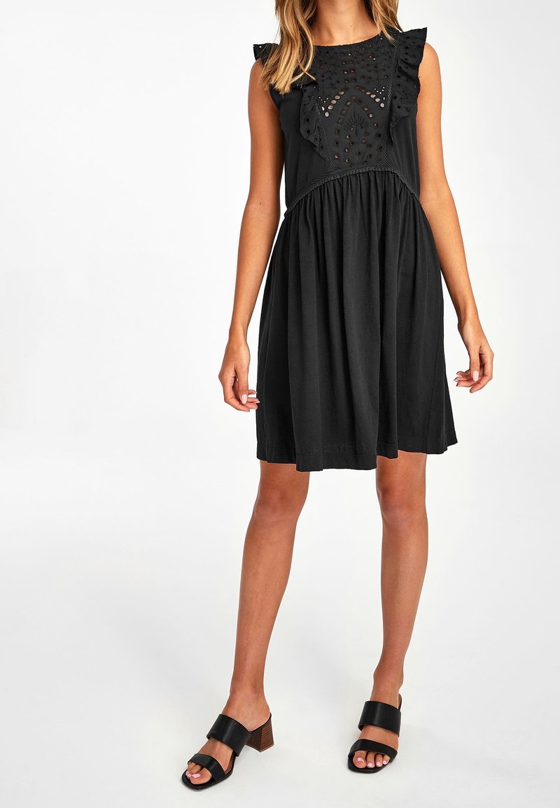 Femme portant une robe noire sans manches avec des détails en œillets et des volants sur la poitrine, associée à des sandales à talons noires à bouts ouverts.