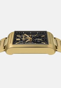 Złoty prostokątny zegarek z przezroczystą czarną tarczą, skomplikowanym mechanizmem i złotą metalową bransoletą. Posiada wyraźne oznaczenia godzin oraz wskazówki.