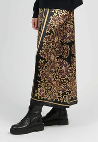 Jupe maxi noire avec un motif paisley doré et bourgogne, présentant une texture soyeuse et une coupe droite, accompagnée de bottes noires à lacets.