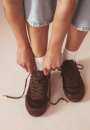 Person mit weißen Socken und hellen Jeansshorts schnürt dunkelbraune Turnschuhe, während sie auf einem hellen Boden sitzt.