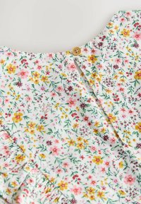 Blumenkleid mit weißem Hintergrund, das farbenfrohe Blumen in Pink, Gelb und Grün zeigt. Auffälliger Holzknopfverschluss am Hals.