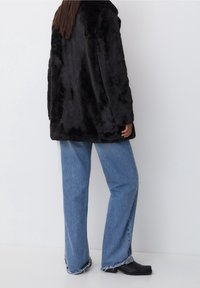 Zwarte nep bontjas met een relaxte pasvorm, iets langere achterkant, gecombineerd met lichtblauwe wijde jeans en zwarte enkellaarsjes.