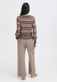Strickpullover mit Zickzackmuster in Dunkelrot, Beige und Braun. Kombiniert mit beigen weiten Hosen, detaillierten Bündchen und hellbraunen Schuhen.