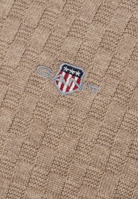 Beżowy sweter robiony na drutach z teksturowanym wzorem. Posiada haftowane logo w kształcie tarczy w kolorze niebieskim, czerwonym i białym z napisem „GANT” pod spodem.