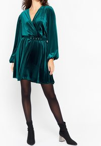 Tealfärgad sammet omslagklänning med långa pufärm, snäv midja med bälte med cirkulär hårdvara och en utsvängd nederdel.