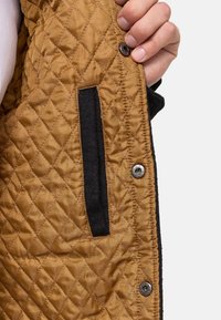 Wnętrze pikowanej kurtki z tkaniną z satyny w złotym kolorze, wyposażone w czarną kieszeń i srebrne zatrzaski. Gładka faktura z wzorem w diamenty.