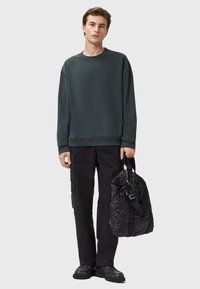 Felpa verde scuro con maniche lunghe, abbinata a pantaloni cargo neri e una borsa a spalla nera e texture. Tessuto morbido, vestibilità casual.