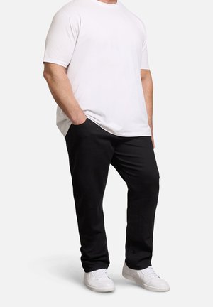 Mężczyzna w prostym białym t-shircie z krótkim rękawem, czarnych spodniach i białych tenisówkach, stojący z jedną ręką w kieszeni na białym tle.