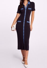 Kvinna i en svart knappradad midiklänning med blåa detaljer och fickor, som håller en vit clutch och bär vita ankelsandaler.