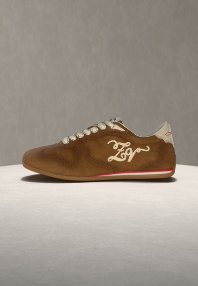 Καφέ σουέντ αθλητικό παπούτσι με λευκά κορδόνια, λευκή μαλακή φτέρνα και λευκή κεντημένη επιγραφή στο πλάι, τοποθετημένο σε φωτεινή στρογγυλή επιφάνεια.