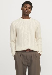 Cream cable knit sweater with a ribbed round neckline, paired with tailored brown trousers featuring pleats and a brown belt.