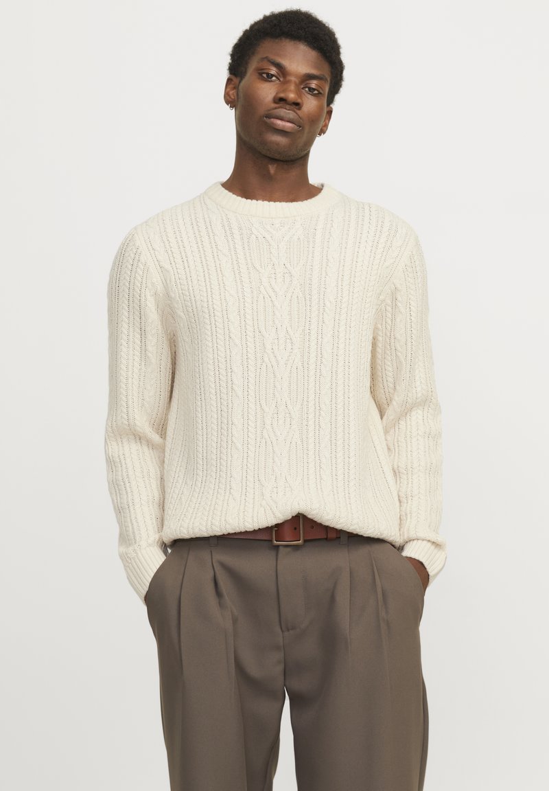 Cream cable knit sweater with a ribbed round neckline, paired with tailored brown trousers featuring pleats and a brown belt.