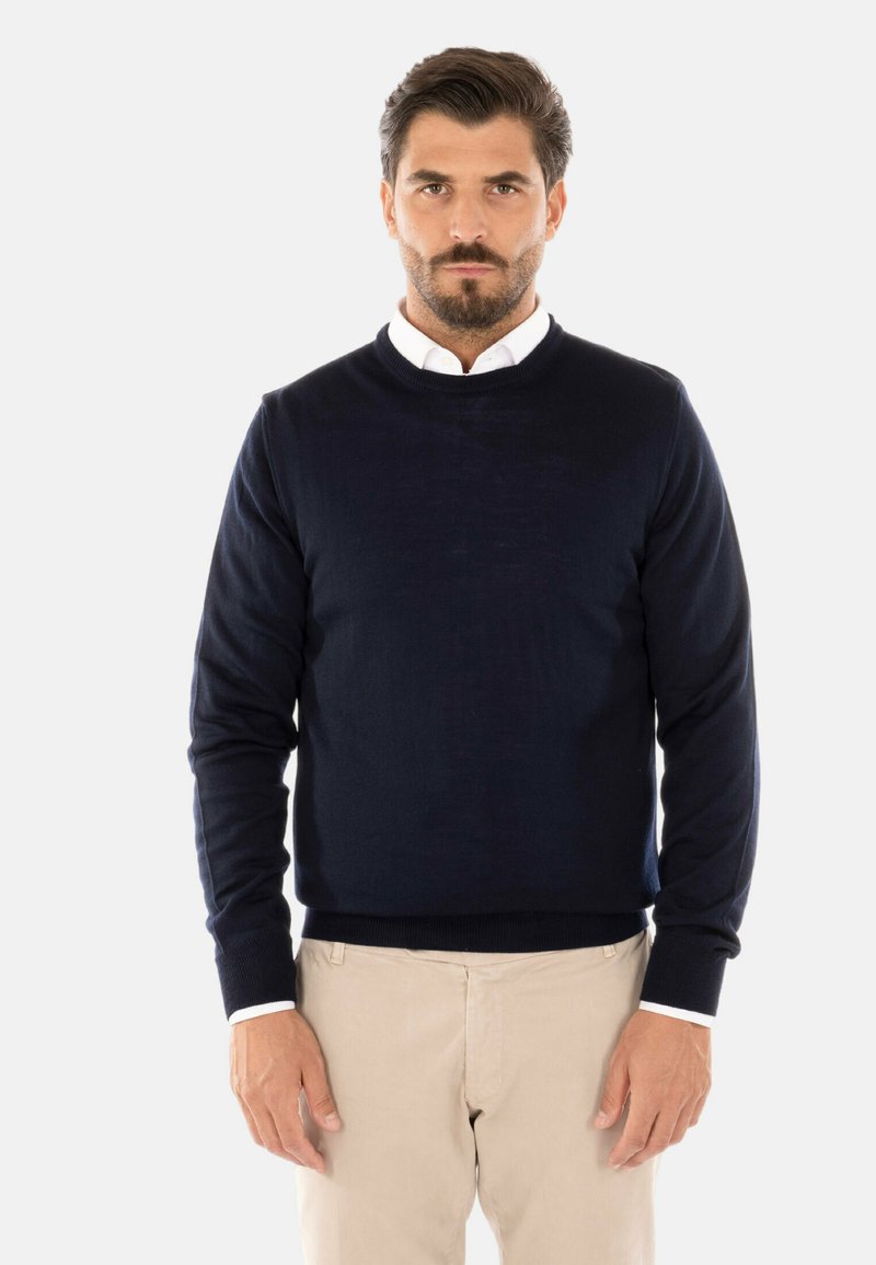 Maglione blu navy a girocollo realizzato in tessuto morbido, con maniche lunghe e polsini a coste, abbinato a una camicia bianca con colletto sotto.