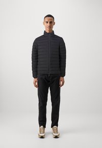 Zwarte pufferjack met horizontaal gestikte secties, hoge kraag en frontale rits, gecombineerd met donkere jeans en beige sneakers.