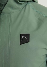 Light green jacket with textured fabric, featuring a black square patch with a white geometric logo and contrasting stitching details.