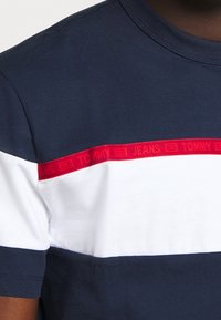 Námorně modré a bílé tričko s červeným vodorovným pruhem přes hruď, které obsahuje nenápadné logo 'Tommy Jeans'.