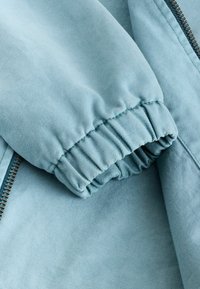 Manche de veste bleu clair avec poignet élastique, couture visible et fermeture éclair en métal partiellement visible sur un tissu texturé.