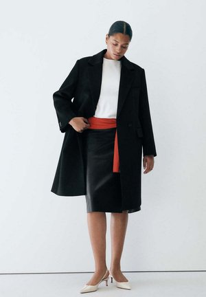 Donna con cappotto nero, top bianco, gonna nera con cintura rossa e tacchi bianchi, in piedi davanti a uno sfondo bianco semplice.