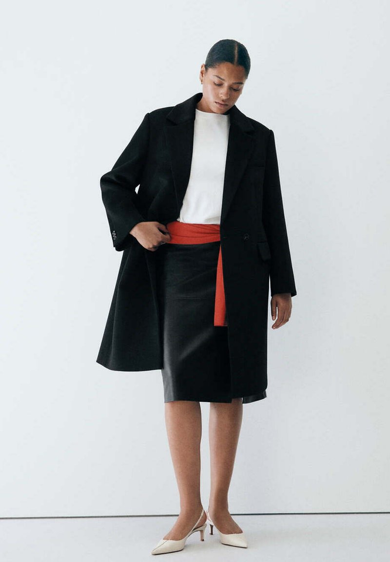Femme portant un manteau noir, un haut blanc, une jupe noire avec une ceinture rouge et des talons blancs, debout devant un fond blanc uni.