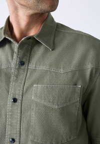 Camisa verde oliva con tejido texturizado, cuello ajustado y botones negros. Presenta un único bolsillo en el pecho con un contorno bordado.