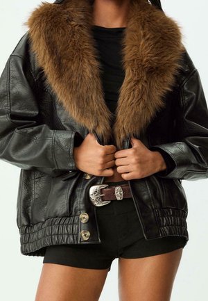 Personne portant une veste en cuir noire avec un grand col en fourrure marron, un short noir et une ceinture marron avec une boucle en argent ornée, les mains ajustant l'avant de la veste.