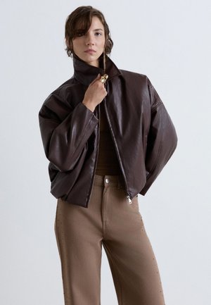 Femme portant une veste en cuir marron foncé oversize, un pantalon beige taille haute, tenant le col de la veste d'une main.