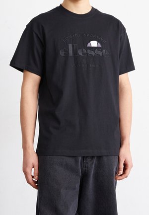 Persona che indossa una t-shirt nera a maniche corte con testo in rilievo "ellesse Italia Lo Stile Sportivo Stabilito Perugia 1969" e logo sul davanti.