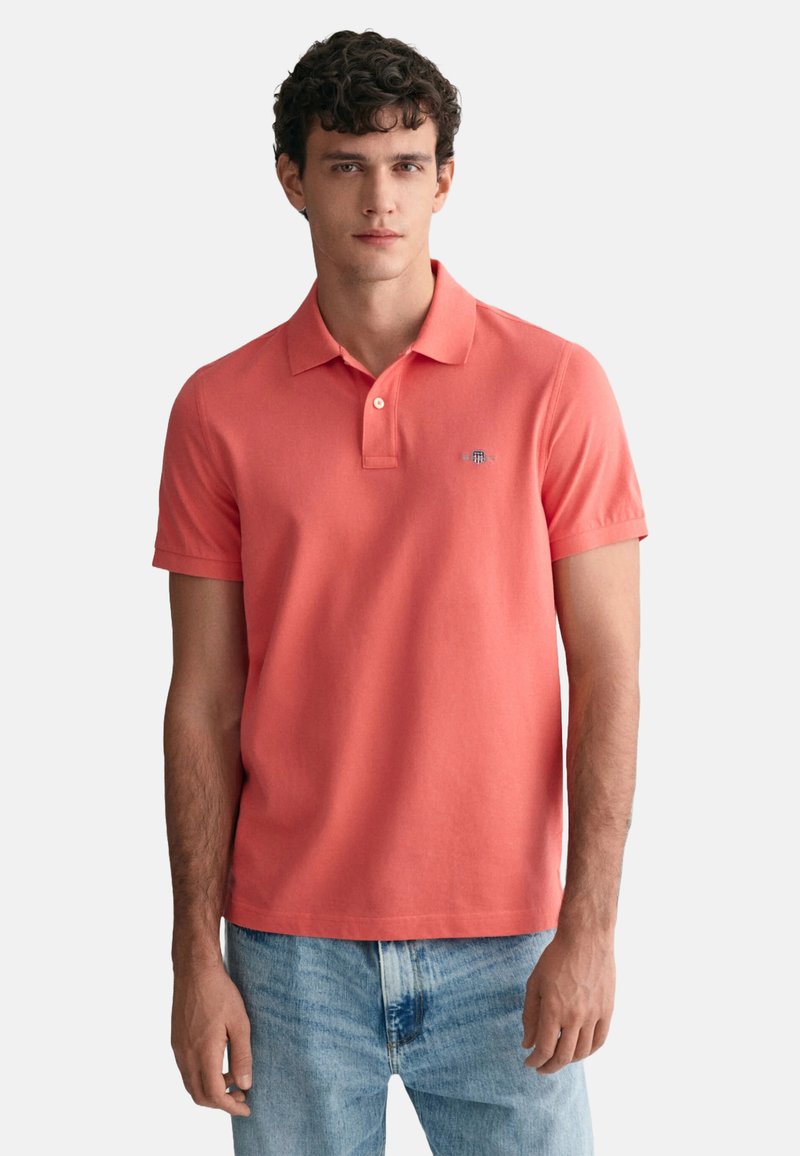 Korallenrotes Poloshirt mit Kragen, kurzen Ärmeln und einem kleinen blauen Logo auf der Brust. Getragen mit hellblauen, ausgewaschenen Jeans.