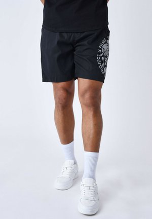 Bañadores negros con una textura suave, que presentan un estampado gráfico en gris contrastante en el lateral. Combinados con zapatillas blancas y calcetines.