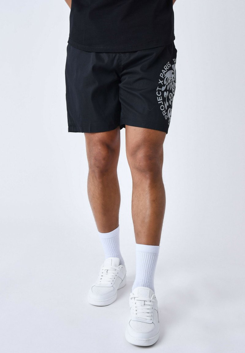 Bañadores negros con una textura suave, que presentan un estampado gráfico en gris contrastante en el lateral. Combinados con zapatillas blancas y calcetines.