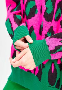 Sweter dzianinowy w różowo-czarny wzór w panterkę, z zielonymi ściągaczami na mankietach i dolnej krawędzi. Zbliżenie na ręce poprawiające rękawy.