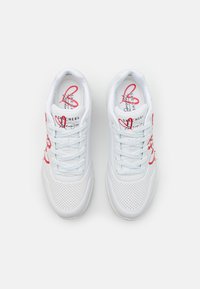 Ett par vita Skechers sportskor med röda hjärtan och vita snören, fotograferade ovanifrån på en enhetlig bakgrund.