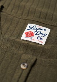 Chemise vert khaki avec un tissu texturé, arborant une étiquette blanche avec un design de rose rouge et un texte bleu, ainsi qu'une fermeture à un bouton.