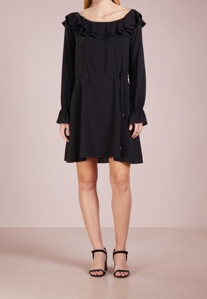 Robe noire décolletée avec encolure volantée, manches longues, taille à cordon et jupe fluide. Associée à des talons noirs à lanières.