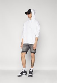 Felpa bianca con cappuccio, pantaloni corti grigi con risvolti, scarpe da ginnastica nere con dettagli bianchi e un cappellino nero. Gli accessori includono occhiali da sole.