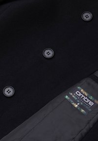 Abrigo de lana negro con cuello muesca, que presenta cuatro botones redondos y una etiqueta interior con un borde de patrón verde. Textura suave.