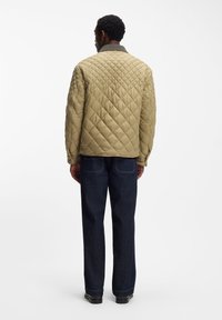 Μπεζ μπουφάν με καπιτονέ σχέδιο και γιακά στέκας, με υφή· συνδυασμένο με σκούρο μπλε τζιν παντελόνι. Η πίσω όψη δείχνει κομψό σχέδιο.