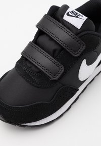 Musta lenkkari, jossa on mokkanahkasta ja tekstiilistä valmistettu yläosa, jossa on kaksi tarranauhaa, valkoinen Nike-logo ja valkoinen kumipohja.